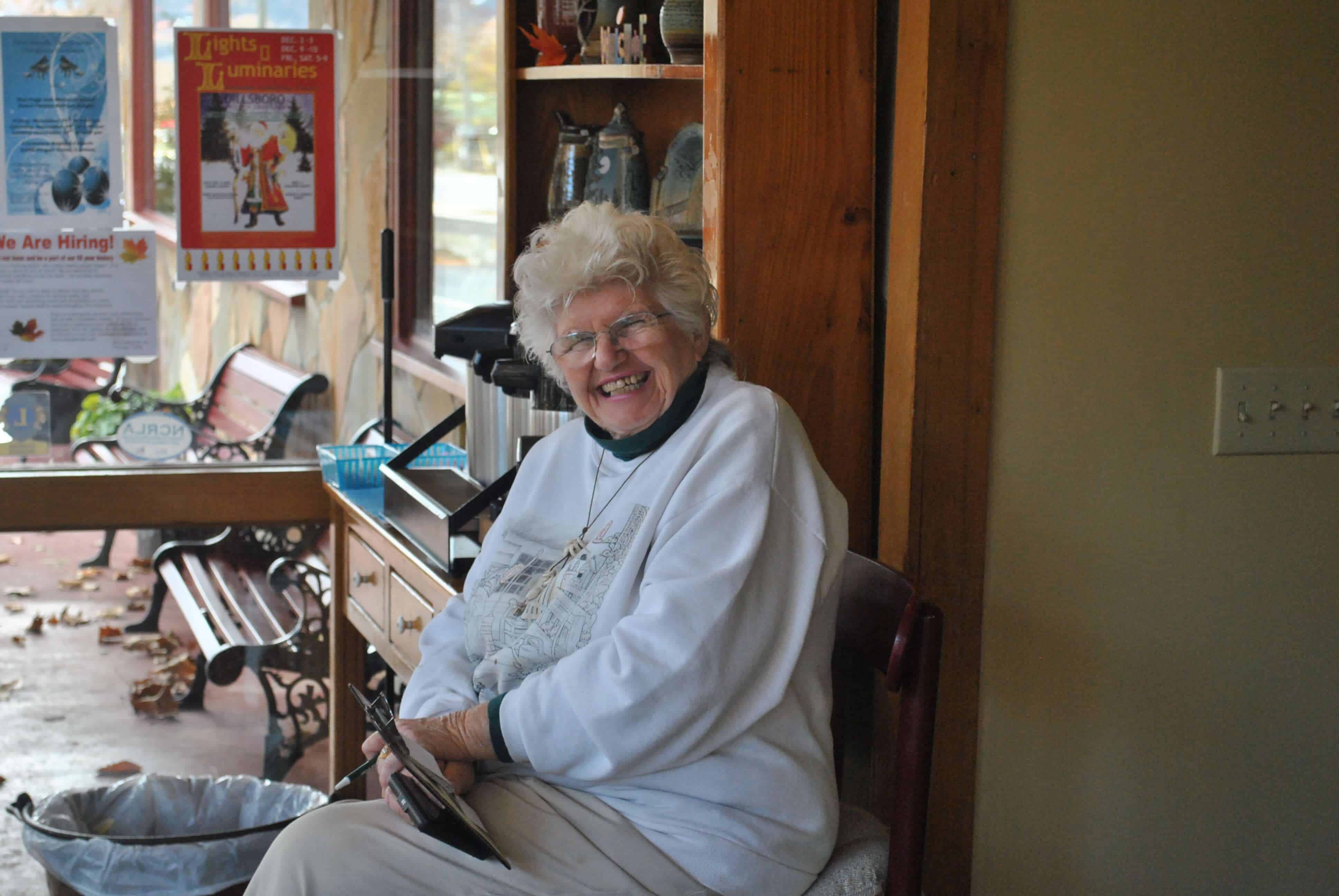 This image shows a lady smiling at the camera. She is located in the front foyer of Joey's pancake house taking peoples request to sit down for breakfast.