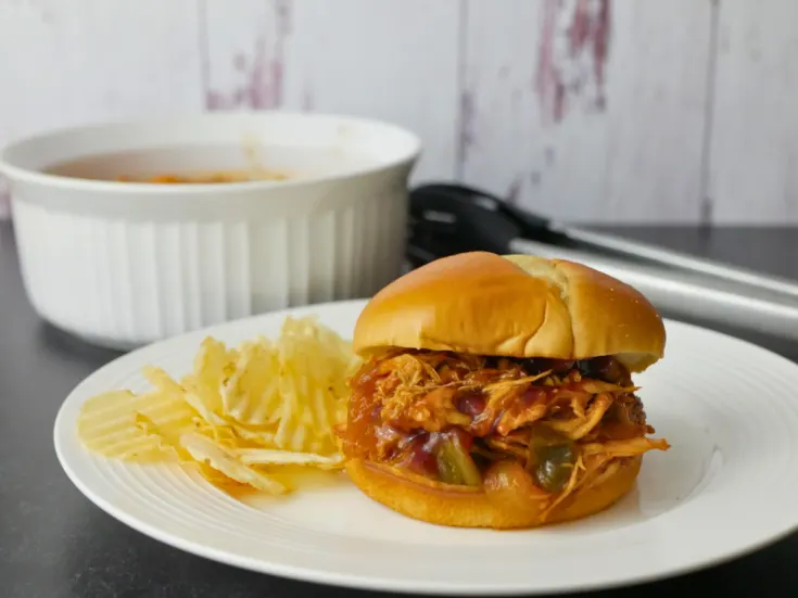 This image is a front view of a sandwich with sweet baby ray's crock pot chicken on a bun sitting on a plate with potato chips behind it.