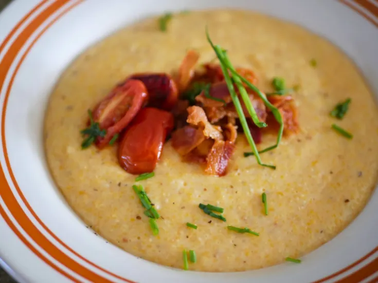 This image shows a bow of cheesy southern cheese grits that is topped with sauteed grape tomatoes, crispy bacon, and chopped chives. The picture is a upclose shot. The bowl is off white with a tan ring around the edge.
