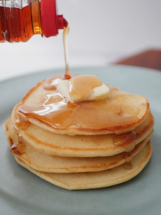 closeup of pouring syrup on a stack of pancakes with butter on top.