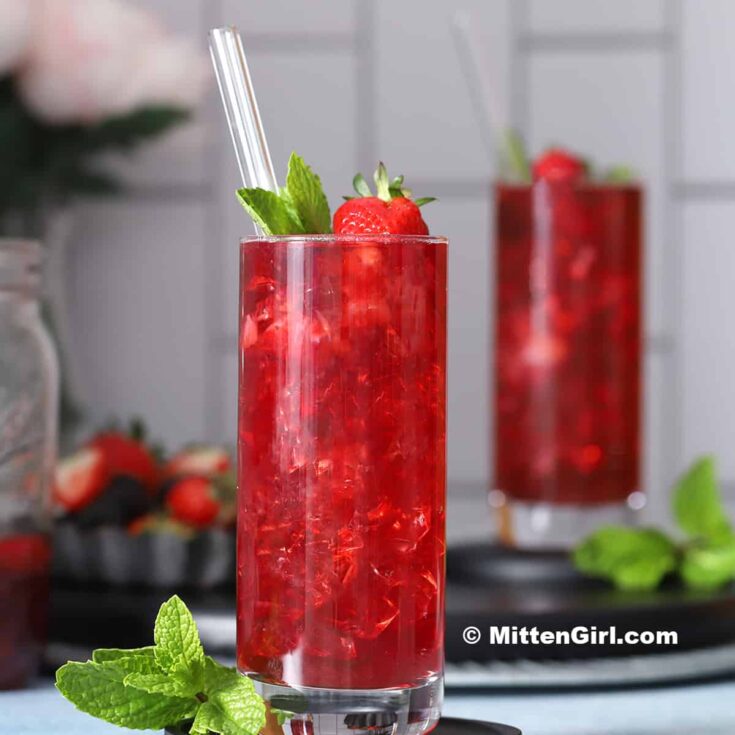 This image is of two tall slender glasses of Berry Hibiscus Iced Tea. Both glasses is full of ice and garnished with a strawberry and mint sprig. Both glasses have clear glass straws sticking out of them.