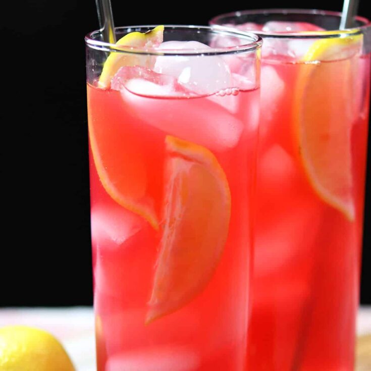 This is a closeup image of two tall slender glasses filled with Hibiscus Lemonade. There is a metal straw in each glass and the drinks are garnished with lemons. The drink itself is a bright pink color.