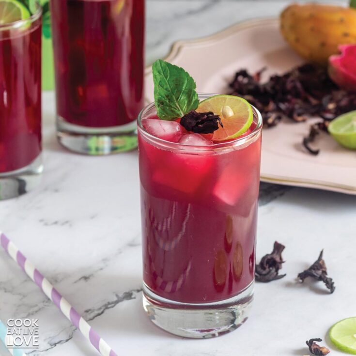 This is a picture of a glass of prickly pear hibiscus agua fresca tea on ice. The color of the tea is bright with a mixture of red and purple. There is a white and purple straw beside the drink on the table and two more glasses full of this tea in the background. The glass is garnished with a mint leaf and Lemmon slice.