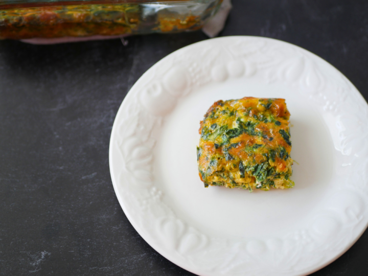 This image is a overhead shot of a slice of Crustless Spinach and Cheese Quiche sitting on a white plate. In the background you can see the rest of this dish in a clear baking casserole dish.