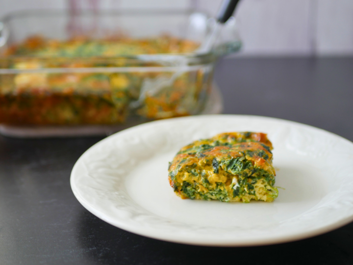 This image shows a slice of Easy Keto Crustless Spinach Quiche on a white plate in the foreground. In the background you can see a baking dish full of this quiche with a slice taken out. There is a black and silver spatula sticking out of the baking dish.
