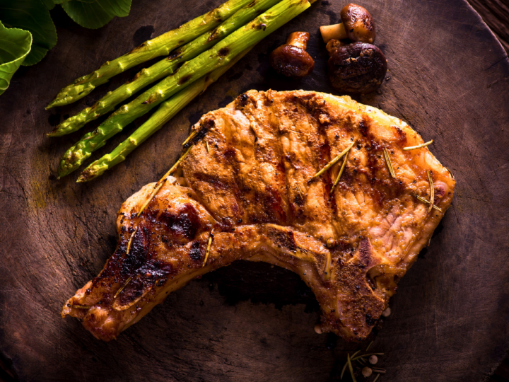 This is a image of a grilled pork chop that is sitting on a dark plate. just above the pork chop is grilled asparagus. 