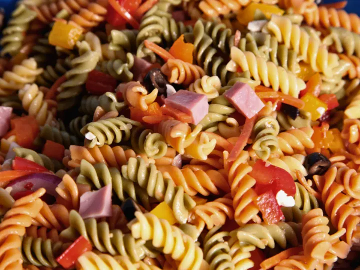 This image is a close up of a classic pasta salad that has spiral noodles of different colors, black olives, cheese, tomatoes, and ham. 