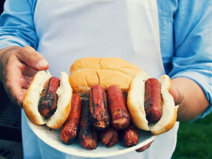 this is a image of a man holding a plate. The plate has grilled hot dogs on it. Some are plain and some are in a bun. The man is wearing a blue shirt with a white apron on. 