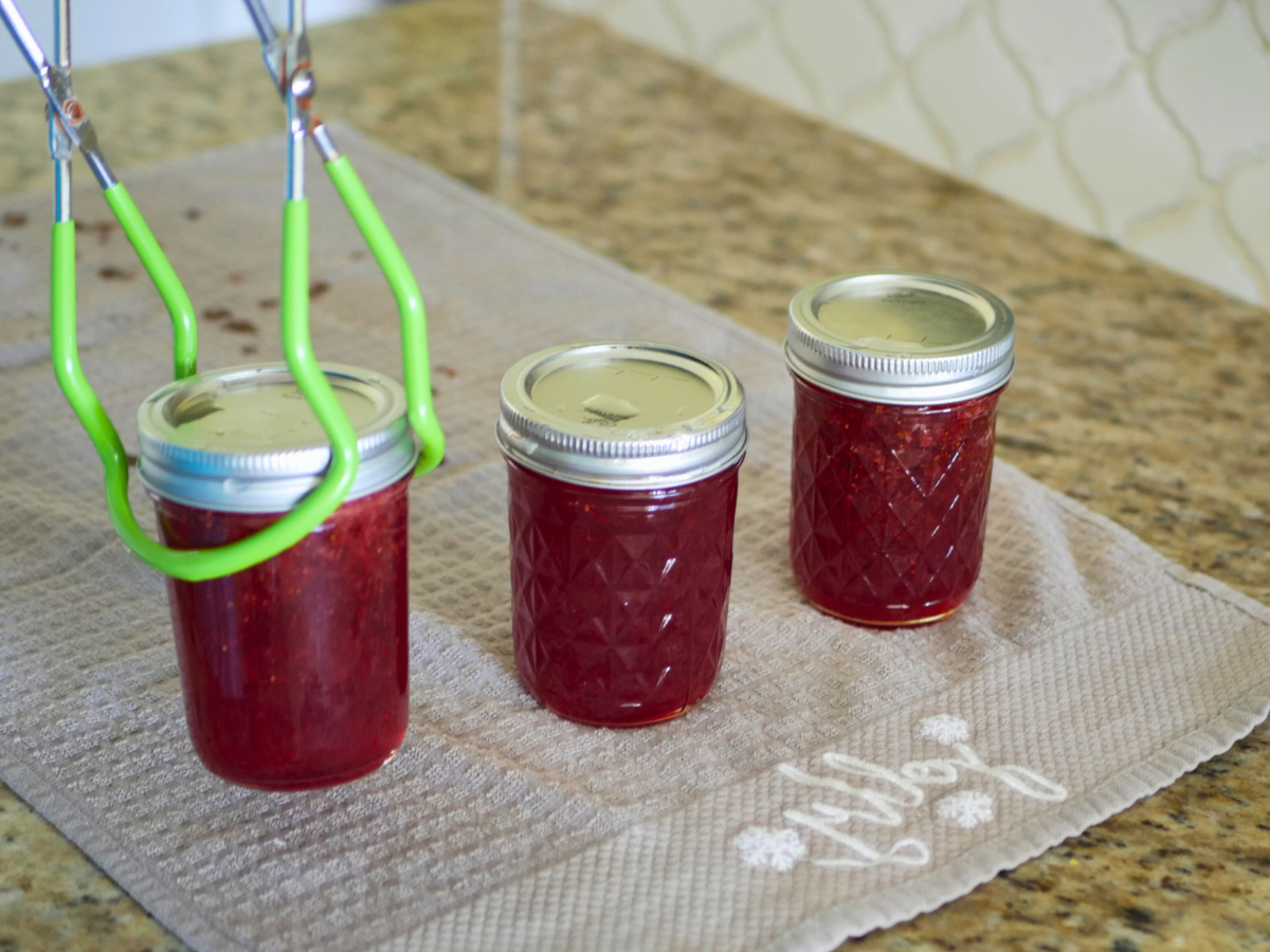 This image shows using a Jar lifter to put Hot Jars of Jam on a tea towel to cool to room temperature after being processed in a water bath canner.