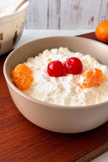 Creamy Southern fruit fluff dessert in a beige bowl topped with mandarin orange slices and maraschino cherries on a wooden table.