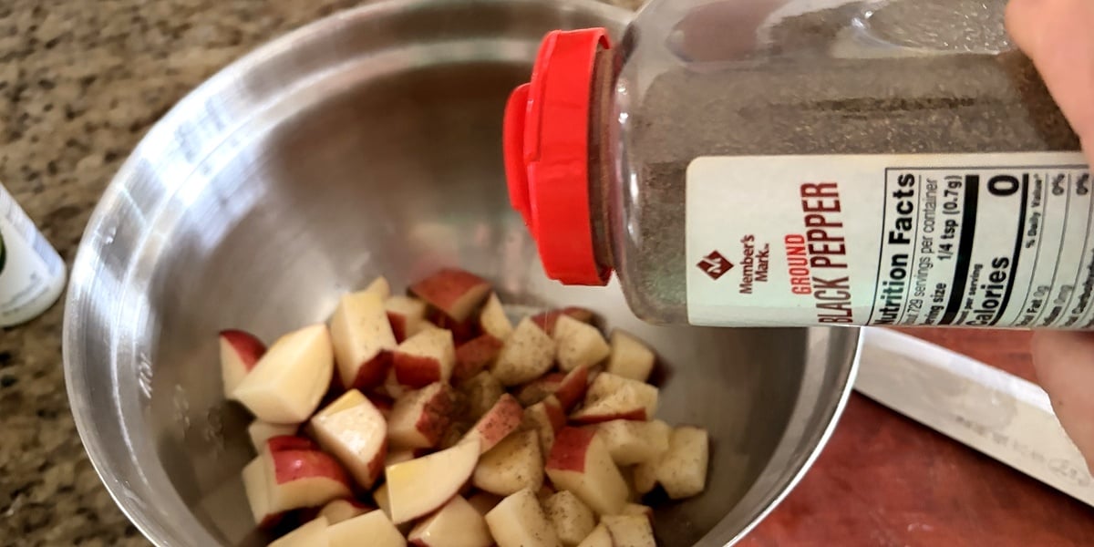 Black pepper being added to diced red potatoes in a mixing bowl before air frying