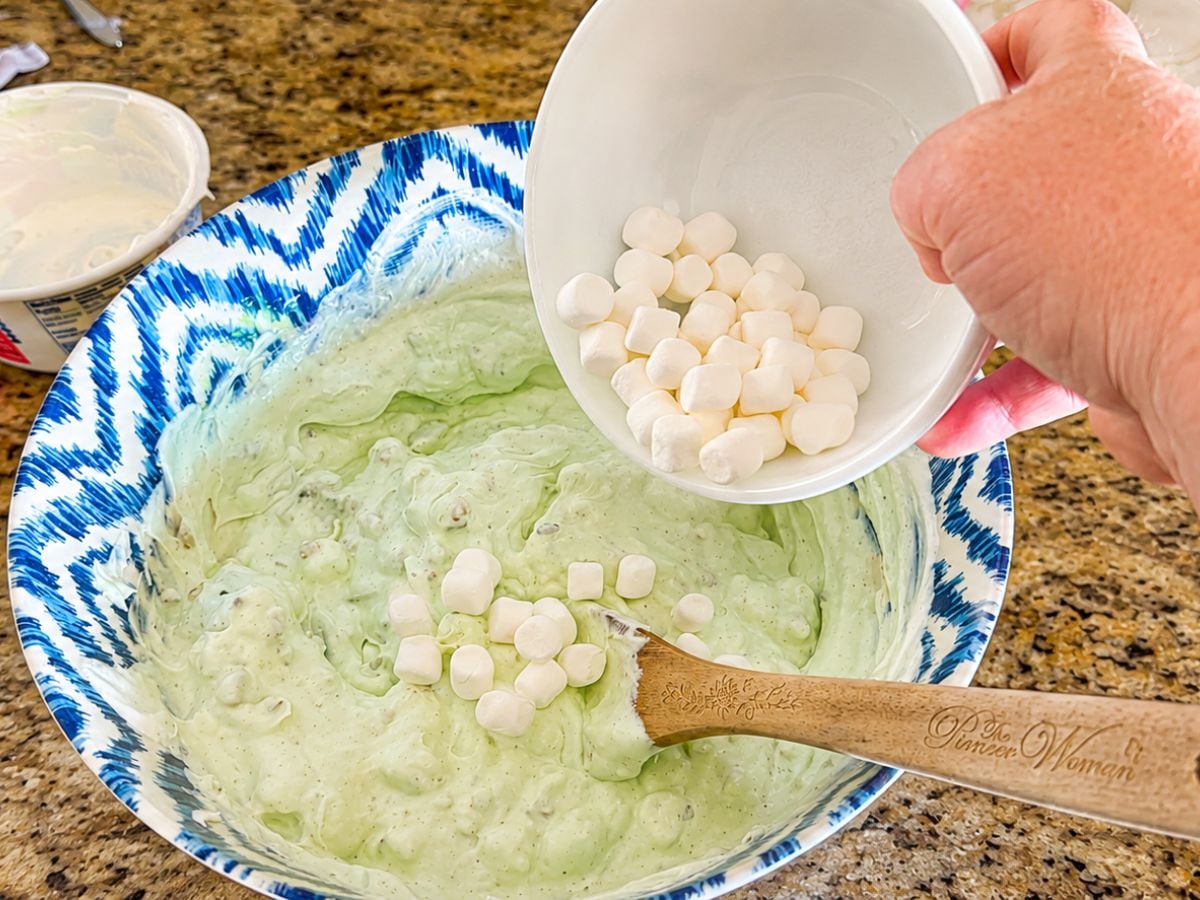 Adding mini marshmallows to the creamy pistachio pudding mixture while making old fashioned Watergate Salad.