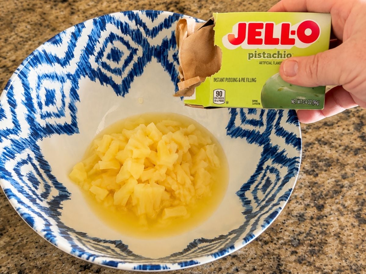 Adding pistachio pudding mix to crushed pineapple and juice in a mixing bowl for Watergate Salad.