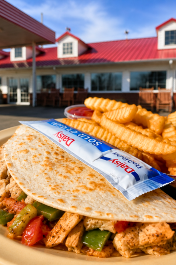 A crispy grilled chicken quesadilla filled with peppers and onions, served with crinkle-cut fries and Daisy sour cream packets on a plate outside Strawberry Hill Cafe in Chesnee, SC.
