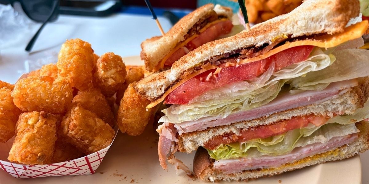 A stacked double-decker club sandwich with turkey, ham, cheese, lettuce, and tomato, served with a side of crispy tater tots at Strawberry Hill USA Cafe in Chesnee, South Carolina.